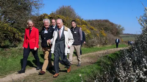 Getty Images King Charles walking along a coastal path on a sunny day yellow gorse bushes in the background. He is joined by 5 other people who are walking with him.