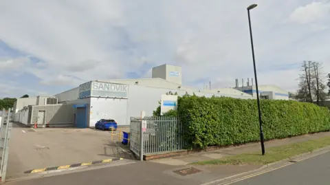 A street view of a large grey factory with blue signs and a hedge in front.