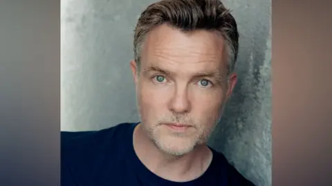 Michael Shelford A man with blue eyes stares into the camera. He is wearing a navy top. He has brown hair swept and style back with shorter grey hairs at the side. He has grey stubble.