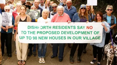 A mixed age and gender group of people standing in front of a countryside field. They are holding up a large sign that reads: "Dishforth residents say no to the proposed development of up to 98 new houses in our village". Others are holding up smaller protest signs.