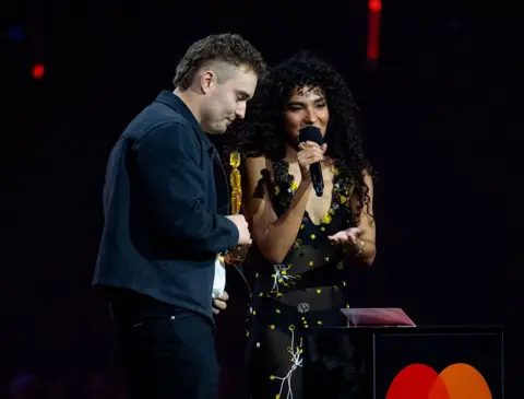 Getty Images Picture of Sam Fender in a blue jacket, holding a Brit award as Olivia Dean speaks into a microphone on stage wearing a black and yellow dress