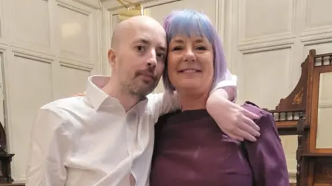 A man and a woman are looking at the camera - the man, who is bald with light facal hair and wearing a white shirt, is on the left. The woman, whose hair is dyed blue and violet, is on the right. She is wearing a burgundy top. In the background are white walls. 