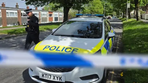 A police officer next to a police car behind blue and white police tape at a cordon set up after a World War Two bomb was found in Plymouth. The area is a residential street with houses on either side of the road. The police tape is blurred.