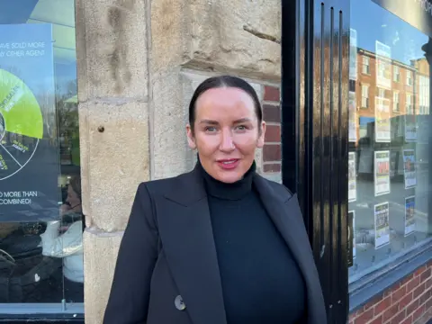 Tammy Daly with long dark brown hair and brown eyes wearing a black polo neck jumper and black blazer sits in an estate agents office. She stands outside an estate agents shop on the corner of a street. A woman can be seen on the telephone in the left window and advert for houses for sale in the window on the right. She is smiling.