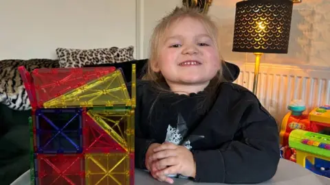 BBC Dani-Rae Brown sat at the table with her elbow on the table. To her right is a multi-colored plastic cube. 