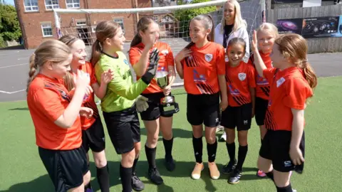 Eight girls wearing orange football tops and black shorts standing in front of a goal. The girls are smiling and some are making high-five gestures, and one holds a trophy. 