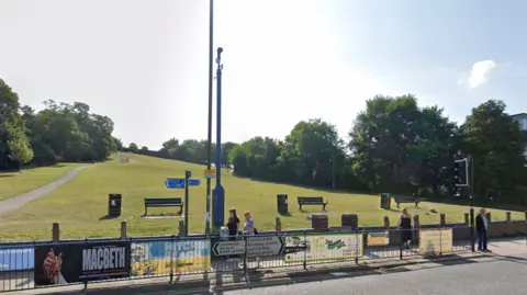 Google A view of a steep hill taken from a road. There are road signs on railings at the bottom of the hill. There is a CCTV camera at the bottom of the hill as well as benches and bins and people walking on the pavement.