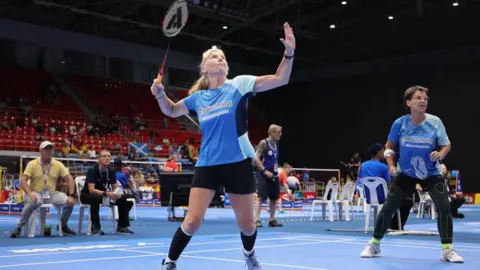 Pamela Peard Both Pam and Sian stand ready for a round of badminton on a blue court. Left is Sian, wearing black shorts and a blue t-shirt. She has a blonde ponytail, holding her racquet up in a serving position. To the right stands Pam who looks focused holding her racquet lower. She is wearing black leggings and a blue t-shirt. Two male coaching staff sit behind the pair on white chairs.