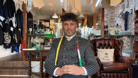 A man with dark, curly hair sitting down in a tailor's shop wearing a pinstriped black and white, doubled-breasted suit with a yellow tape measure around his neck.