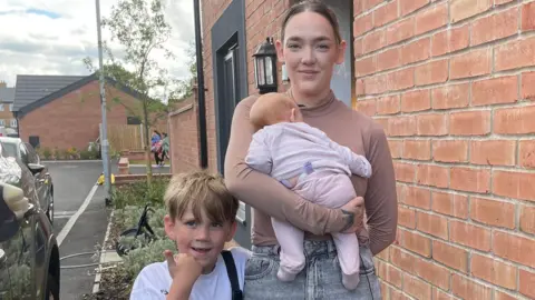 Lauren wearing a pink top holding a baby in her arm, with Austin beside her outside a red-brick property