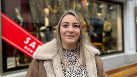 The image shows Rute Dos Reis, in a brown jacket and a leopard print shirt, smiling at the camera. She is standing outside, in front of a shopfront window.