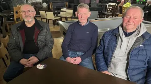 BBC Three men are sitting on chairs inside a pub ahead of their interview. There is a pool table, a bar and tables and chairs in the background. 