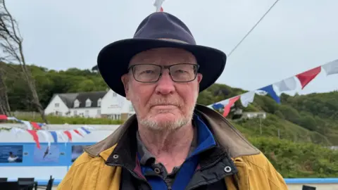 Andrew Le Quesne is wearing a black hat, yellow jacket and glasses. 