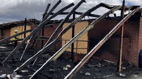Shaun Whitmore/BBC A row of planks of wood that have been damaged and blackened by fire lean at a 45-degree angle against a red brick building which had no roof. The ground is covered in soot and blackened detritus from the fire. The sky is cloudy. A man in a hi-vis jacket and white hard hat looks at the building. 
