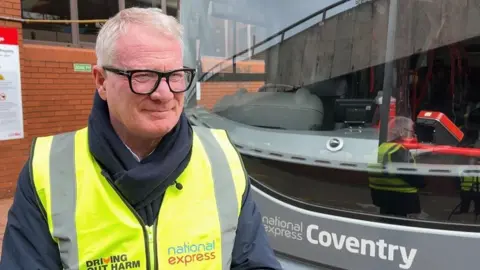 Richard Parker is standing in front of an electric National Express Coventry bus. He is wearing a yellow hi-viz vest over a blue waterproof jacket