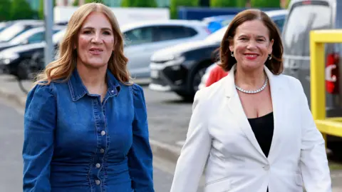 PA Media Janice Boylan, left, with long auburn hair, wearing a denim long sleeve top. She is standing beside Mary Lou McDonald, with shoulder length brown hair, wearing a black top and white blazer. 