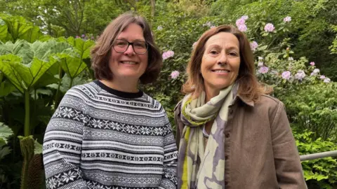 Alison Marshall is on the left of the photo wearing a black and white jumper. Melina Syred is on the right of the photo with a brown blazer and a green and purple scarf. They are pictures in the Vallee Des Vaux peace garden. 