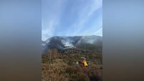 Derbyshire Fire and Rescue An area of steeply sloping moorland from which a quantity of smoke is rising and a firefighter is visible in the foreground