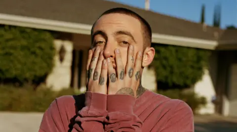 Mac Miller poses with his hands on his face for a magazine shoot. He's pictured outside a single-storey house on a blue-sky day. He has croppped brown hair, brown eyes and tattooed fingers and wears a red sweater. 