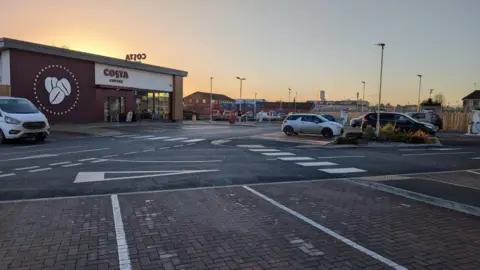 A car park outside a Costa drive-thru coffee shop with a large coffee beans logo on the site. A van and several cars are parked outside. It is dusk and the sky is orange and blue.