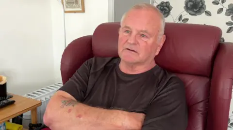 77-year-old Elvet Jones sitting in a maroon leather armchair, with a brown t-shirt and his arms folded