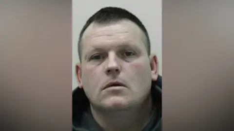 Northumbria Police Mugshot of David McNeil. He has pale pasty skin and a large clean shaven face with short black hair shaved at the sides