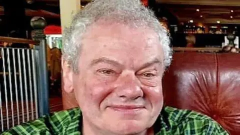 Family photo Brad smiling and wearing a green plaid shirt sitting in a leather chair. A bar can be seen behind him. He has short silver curly hair