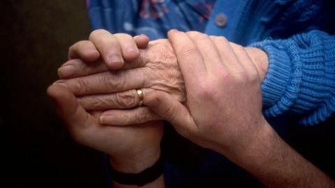 A pair of hands clasped around the hand of someone wearing a blue knitted cardigan and a gold wedding ring
