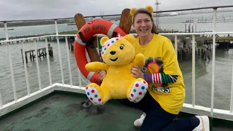 Katie Martin in all yellow and Pudsey ears holding a Pudsey bear soft toy as she travels on the ferry to the Isle of Wight.