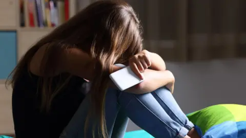 Getty A young woman with long dark hair sits on a bed with her head in her arms. She is holding a white smartphone and is wearing a dark sleeveless top and light blue jeans.