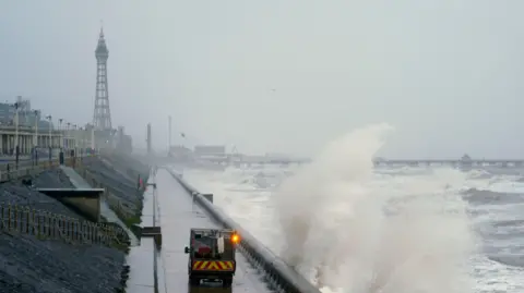 PA Media The spray of large waves rise as they crash into the coast of Blackpool with Blackpool Tower rising up behind on the left on a grey day. A maintenance vehicle is driven on a road next to the waves. The pier with a ferris wheel can be seen stretching over the water behind.