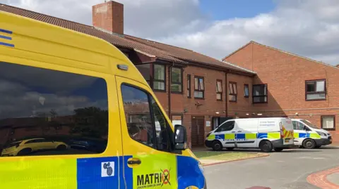 A yellow police van and two white ones parked outside a two-storey red brick retirement home. One of the vans has crime scene investigation written on the side