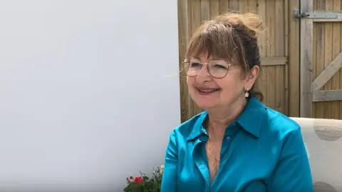 Zoe Cousens is sitting on a chair outdoors. There is a whitewashed wall and a gate beside her. She is wearing a blue silk blouse and her brown hair is tied up. She is wearing glasses and earrings and she is smiling broadly.