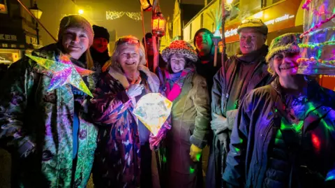 Paul Campbell A group of people, wrapped in outdoor winter clothing, standing in the dark with bright lights lighting them up.