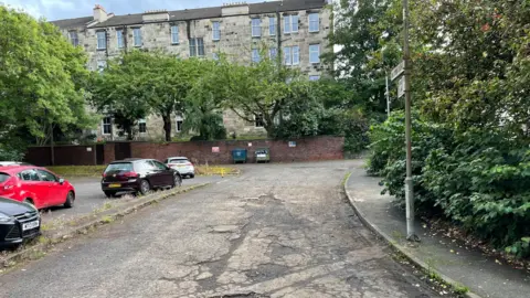 Potholed road with cars parked next to it. There are flats, trees and a street sign and two bins against a wall in the distance 