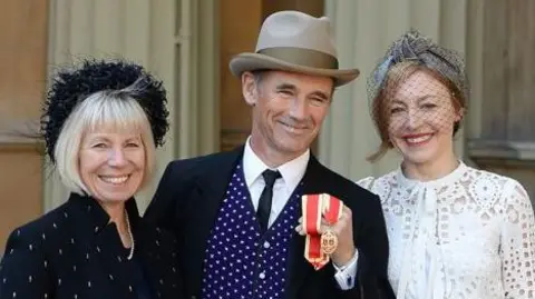 PA Media Claire wears a black headband and a black embroided jacket with a string of pearls as she stands next to her husband Sir Mark outside Buckingham Palace after he was knighted 