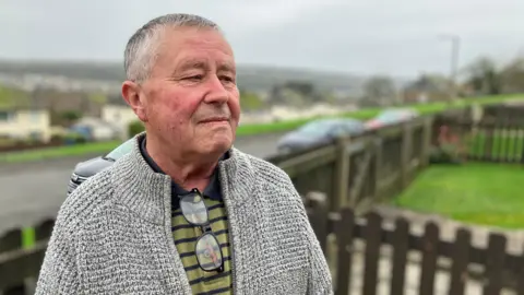 Dave Cradduck stands in a garden overlooking the town of Whitehaven. He has a stern expression and he is looking at something out of shot. He has short grey hair and is wearing a grey woollen cardigan over a green and blue polo shirt. A pair of glasses are hanging from the collar of his top. 