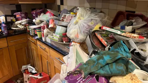 Adrian Harms/BBC A close up of the rubbish pilled up in the kitchen of Nevena Khediri's home in Camberley, Surrey. Food boxes, sweet boxes and filled plastic bags can be seen on the kitchen surface.