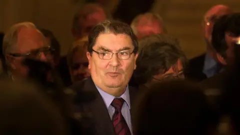 Pacemaker John Hume stands among a crowd of people. He is wearing glasses and has short dark hair. He is wearing a suit, shirt and tie