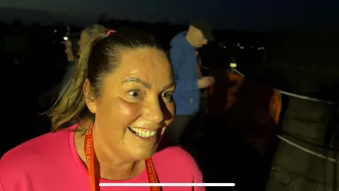 Donna smiling to the right of the camera. She has brown hair which is tied back in a ponytail and is wearing a bright pink t-shirt with a red medal around her neck. It is dark outside and he looks slightly flushed. There are people standing behind her.