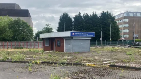 An old bus station in Wythenshawe, situated on an overgrown car park. The bus station is shut up with a corrugated iron shutter, the rest of the building is made of dark red brick and has a flat roof.