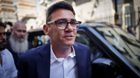 Andy Burnham is walking alongside a parked vehicle. He is wearing a blue jacket, a white shirt and glasses. 