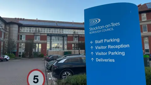 A blue sign stands to the right which reads in white lettering, Stockton-on-Tees Borough Council, staff parking, visitor reception, visitor parking and deliveries. A large brick building called Dunedin House and car park sits behind it. 