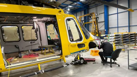 A man wearing black overalls is sitting on a wheelie chair and leaning towards the nose of the yellow helicopter which he is working on in a hangar. The helicopter's door is open and there are tools and a small stool inside it.