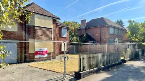 Two large houses with driveways. The windows of both houses are bordered up and the gardens are surrounded by metal fencing. There is a sign on the fencing that reads "KEEP OUT."