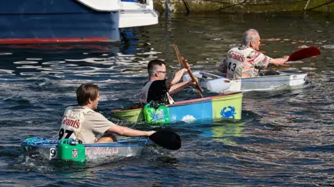 MANX SCENES Three competitors in a row paddling tin baths. They are each wearing a bib with a competitor number on the back.