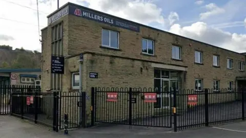 A yellow stone factory surrounded by black iron gates with a 'Millers Oils' sign