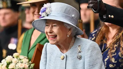 Queen Elizabeth II wearing a powder blue jacket and hat, with white and lilac flowers on the hat, is looking to the left and smiling broadly 