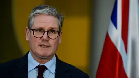 PA Media Prime Minister Sir Keir Starmer looks at the camera. He is wearing glasses, a dark suit and tie and blue shirt, with a flag in the background on the right side of the photograph. 