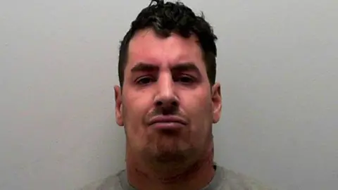 A man with black hair, big black eyebrows, a goatee and pink face frowns at the camera. 